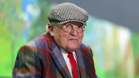 Getty Images British painter David Hockney poses at the Orangerie museum in Paris, on October 7, 2021, in front of his painting "A year in Normandy", a 91-meter-long artwork painted during the lockdown in 2020. - Hockney made a hundred drawings on iPad in a matter of weeks and captured the effects of light and climate change during the four seasons.