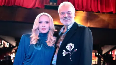 Ricardo Gomes Madonna and Graham Norton are pictured together. She is wearing a blue dress, he is in a dark jacket with a flower motif. They are photographed against a nightclub background.