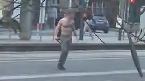 UGC A still from a video showing a bare-chested man wearing dark trousers standing in the middle of a street. He is carrying a weapon in his hand. The still is blurry and the man's face has been delibrately blurred.
