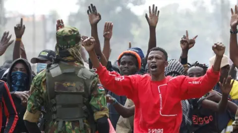 Reuters A group of protesters - one in a red top clearly seen at the front - hold their arms in the air as they face a policeman in camouflage uniform.