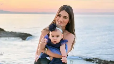 Instagram A woman with long dark hair smiles as she holds a toddler in a dress on a beach, the sunset behind her