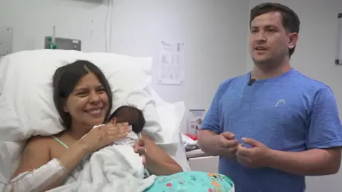 Dayana Patino grins as she holds her son Juan David close to her chest from a hospital bed. Her husband, Gerson, stands beside them wearing a light blue t-shirt. 