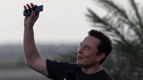 Reuters Elon Musk wearing a black T-shirt and holding his phone above his head.
