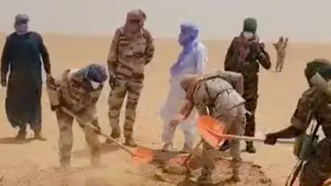 Agadez governorate/Facebook People, including several in military-style uniforms, use shovels to dig in a sandy desert area, with dust rising around them
