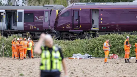EPA/Shutterstock Men in orange hi-vis and white hard hats look at two trains, which have crashed head-on into each other. The trains are dark purple. In the blurred foreground a police officer walks away from the scene, towards the camera.