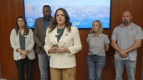 City of Midland A woman in a white blazer stands in the foreground with a cell phone in her hands as she speaks. In the background of her are four other individuals leaning up against a wooden wall. 