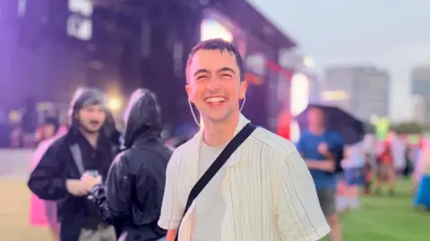 Alex Milsom Alex Milsom is wearing a wet striped t-shirt and has wet hair. He is standing in a festival area in front of a stage with other people wearing rainproof jackets.