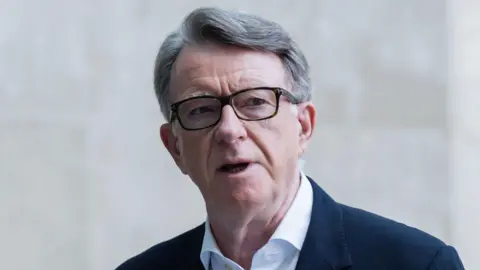 Getty Images Lord Mandelson wears glasses, a white shirt and blue suit jacket, he is standing outside a stone building 