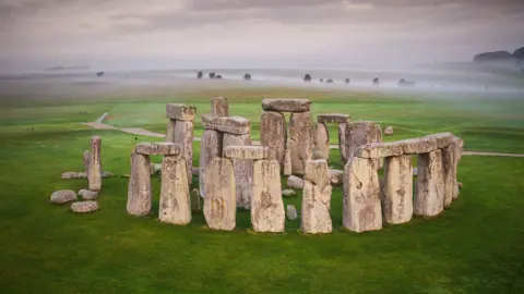English Heritage The image shows Stonehenge. There is a misty sky behind with the full circles of the ancient monument in the foreground. Four sets of two large upright stones, each with another large stone resting across the top, which make up part of the outer circle.  The even larger stones of the inner area are visible above those of the outer circle. Green grass surrounds the monument on all sides.  