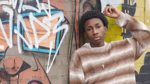 Ziame Stewart A boy with black curly hair poses against a graffitied wall