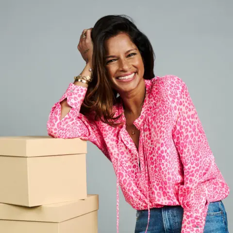 BBC Dilly wearing a pink patterned long-sleeve top leaning on a stack of beige boxes in the foreground, with one hand resting against the side of their head. Several bracelets, rings, and a necklace are visible. The background is a plain light grey wall.