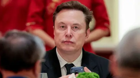 Reuters Elon Musk seated holding an iPhone at an event wearing a suit and tie.