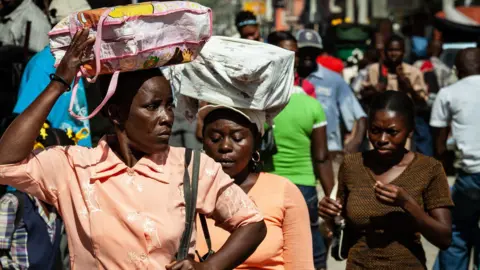 Getty Images Three Haitian women carry bags on their heads as they walk down a crowded street. Two women wear orange blouses and one wears a brown t-shirt.  