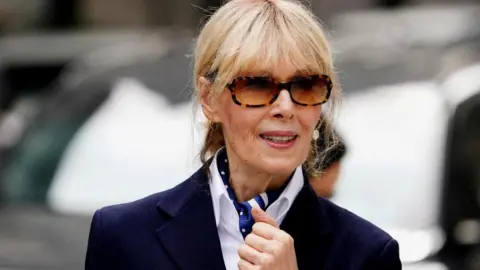 Reuters E Jean Carroll with a navy blazer, white shirt and blue patterned scarf. She is wearing large sunglasses and looking straight ahead. 