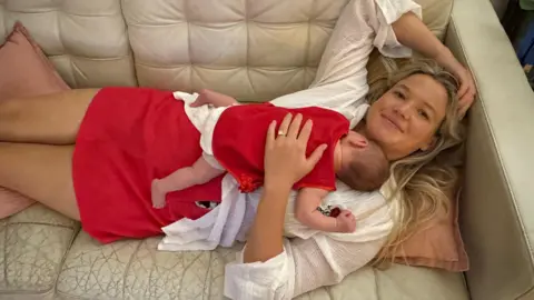 Supplied A woman with long blonde hair, wearing a white top and red skirt lying on a cream couch, with a baby sleeping on her chest. 