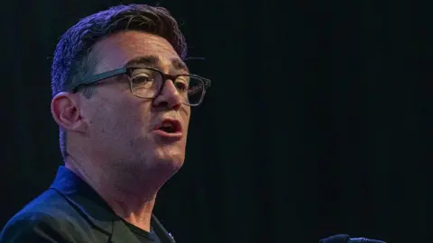 Getty Images Andy Burnham wearing a black jacket with a black t-shirt talking to an audience with a black background behind him