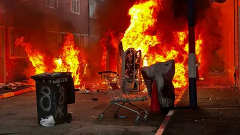 PA Media A wheelie bin and a trolley and a car are all on fire on a street. The kerb stones are painted red, white and blue.