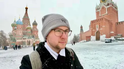 Ben/ BBC Person in the foreground wearing a grey beanie hat, glasses, a dark coat and a backpack, with snow on clothing and shoulders. Snow-covered cobbled square surrounds the scene. In the background, St Basil’s Cathedral with colourful onion domes stands to the left, and a red brick Kremlin wall with a tower is on the right. Several pedestrians walk across the square, and a parked vehicle is visible near the wall. Snow is falling lightly across the image.