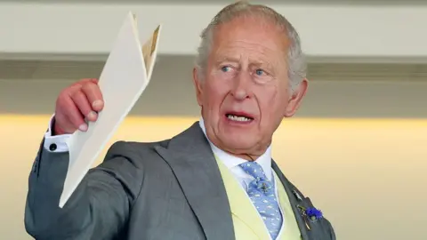Getty Images King Charles holding a white A4 booklet and gesturing with it. He is wearing a pale great suit, cream waistcoat, blue and white patterned tie, and a white shirt.