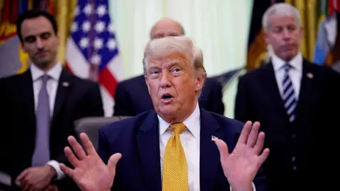 Getty Images US President Donald Trump wearing a navy suit, white shirt, and gold tie holds both of his hands up as he speaks during an event in the Oval Office of the White House. Two mean in suits and ties are on located on the left and right of him.
