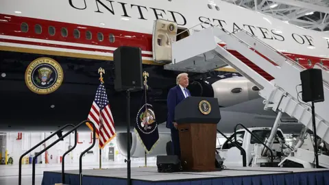 Getty Images Trump standing at a podium on a stage in front of the new jet, painted white with dark red and blue stripes. There is a presidential seal and the words "United States of" visible. 