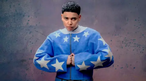 Michelle Helena Janssen The musician KWN is pictured in a "strongman" pose, with her hands pressed together in fists. She is wearing a blue zip-up jacket with large white stars on the sleeves and collar. 