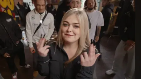 Steeze Factory Morgan Da Savage has blonde long hair and is wearing a black top. She has her fingers up in a "gunfinger" position and smiles into the camera as she performs her rap in the Steeze Factory in front of a group of other people.