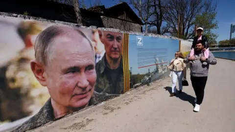 Reuters A poster showing Russian President on the side of a road where a man wearing black trousers, a gret top is carrying a child on his neck, with a woman walking by his side, wearing light blue trousers,a  white top and a pink jacket, as well as carrying a small black handbag 