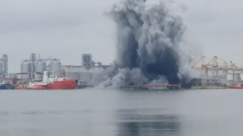 Observator Antena1 A large cloud of dark smoke is seen from a distance rising from a port. Boats, cranes and silos can be seen around it.
