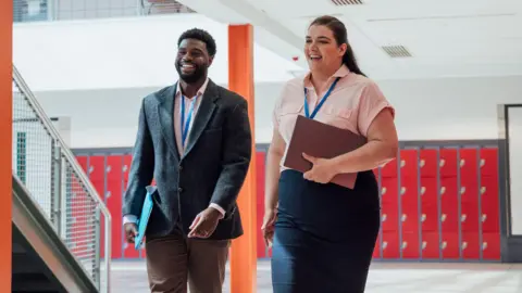 Getty Images/SolStock Two employees walk together while smiling. They're both wearing lanyards and holding folders.  