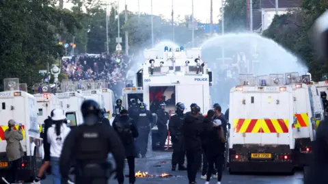 Pacemaker A number of police vans and police in riot car in the middle of a road with the remains of a fire beside one the vans. There is a water cannon near the front spraying a large crowd of people.