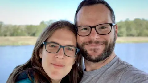 Alan Donegan Katie and Alan Donegan smile at the camera while both wearing glasses during a selfie in front of a lake and trees against a blue sky.