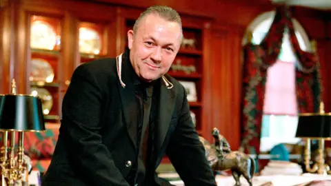 BBC David Sullivan wears a black suit while sitting on a desk in his home in 2003. 