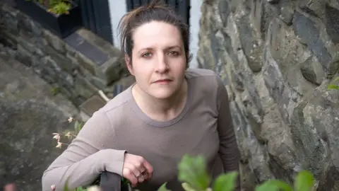 BBC Ruth has dark hair and wears a long-sleeved top, she is leaning on a railing and looking up along a narrow outdoor path, next to a stone wall.
