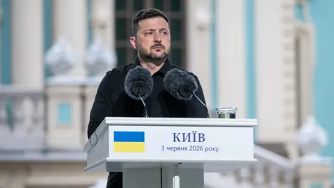 Danylo Antoniuk / Anadolu via Getty Images Zelenksy looks pensive as he focuses his attention to the right of frame while he stands at a podium against a light blue bokeh background, on the podium are two microphones, a glass of water and Ukrainian lettering indicating the date of 3 June alongside a depiction of the Ukrainian flag.
