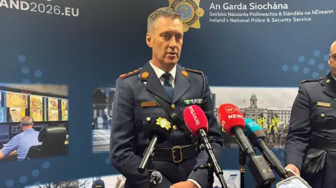 PA Media Garda officer wearing a blue uniform. He has short dark and grey hair and has mics in front of him. An Garda Siochana is on the background behind with police pictures.