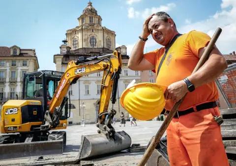 Tino Romano / EPA A construction worker wearing orange hi vis uniform holds a yellow construction helmet and wipes his head in front of a JCB digger next to a public square in Turin, Italy on 25 June.