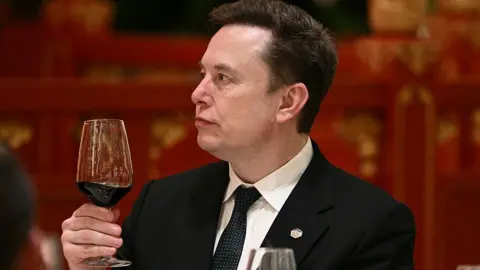 AFP via Getty Images Musk, wearing a black suit, white shirt and black and white dotted tie, holds a glass of red wine while at an event in Beijing, during a recent US trade visit.