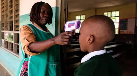 Getty Images A woman in a green tabard uses a thermometer gun to check the temperature of a school pupil who has their back to the camera.