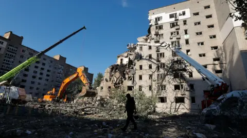 SERGEY DOLZHENKO/EPA/Shutterstock A nine-storey block - the top left corner is missing, and a large digger moving debris 