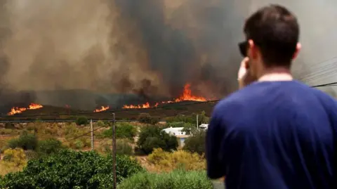 Reuters Flames and black smoke are seen rising along the horizon from a hilly area in the background, while a man in the foreground looks towards it with his hand to his mouth 