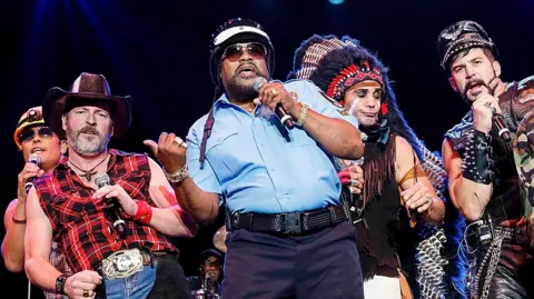 Getty Images Willis in a police uniform singing and dancing at the front of the group in their different costumes, pictured in 2018