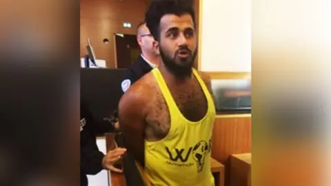 Twana Jamal in a yellow weightlifter's vest with a black graphic stands in an indoor public setting with hands held behind the back, while another individual in dark clothing stands close behind; wood-panelled walls and a counter are visible in the background, and partial text reading “W” appears on the shirt.
