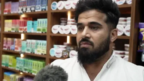 Close-up of Twana Jamal with dark hair and a full beard, wearing a white collared shirt, standing indoors in front of wooden shelves filled with neatly arranged, colourful product boxes and small round containers. A microphone with a fuzzy windscreen is positioned in the lower foreground, indicating an interview or recording. The background features warm lighting within the shelving units, highlighting the rows of packaged items.
