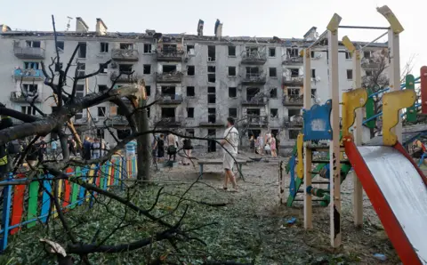 EPA A partially-damaged playground slide in yellow, red and green stands on the right of the frame. A tree split at the trunk is bent over a fence also painted in red, green, yellow and blue. In the background tens of residents walk around the while a partially-destroyed building sits in the background.