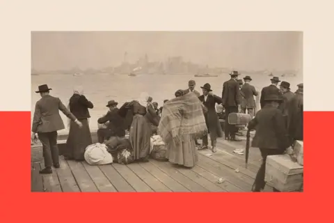 Getty Images A group of immigrants wait at Ellis Island to be taken to Manhattan. They wear cloaks, hats and coats.