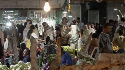 Jack Garland / BBC A busy market scene with lots of people looking and stalls selling vegetables. The stall front left appears to be piled with white cabbages and a man and a woman are sitting behind them. 