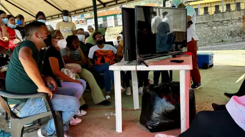 BBC Mundo Families try to identify their loved ones through images scrolling across two screens. Some are sitting in chairs, their faces filled with anguish.