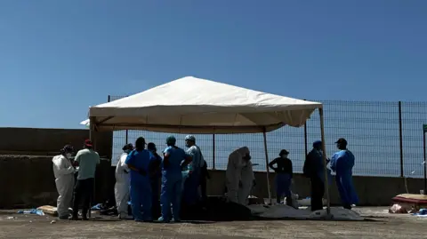 BBC Mundo Medical personnel around bodies on the ground covered with plastic bags. The workers are wearing blue or white gowns.