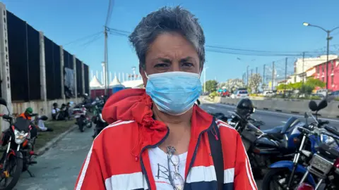 BBC Mundo Modesta Alemán, wearing a blue face mask, stands in front of Los Silos. She is wearing a red jacket with blue and white stripes.

